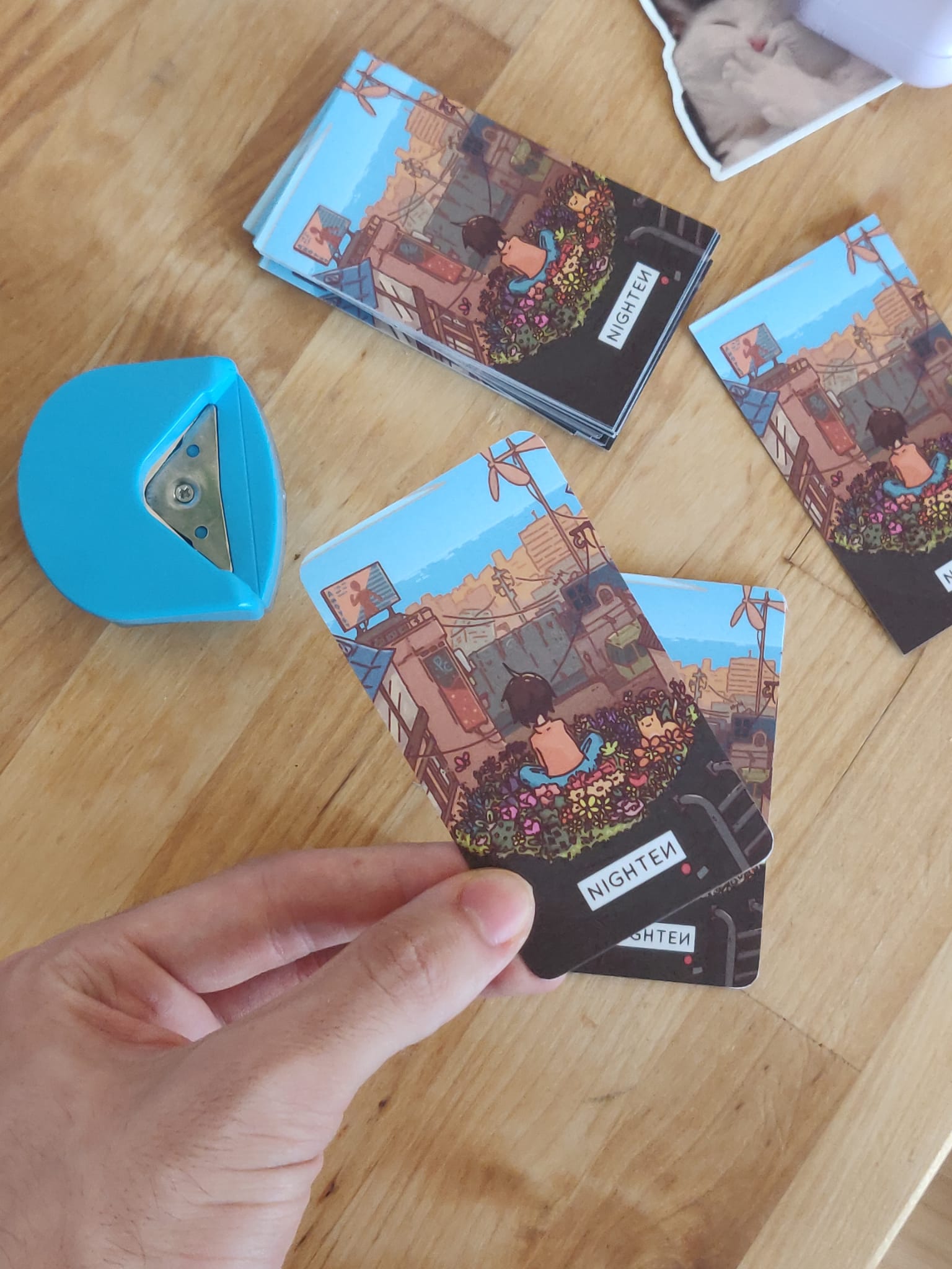 My hand holding a business card with a illustration on it. The corners have been rounded by a little device next to it. A stack of individual business cards is displayed next to it.