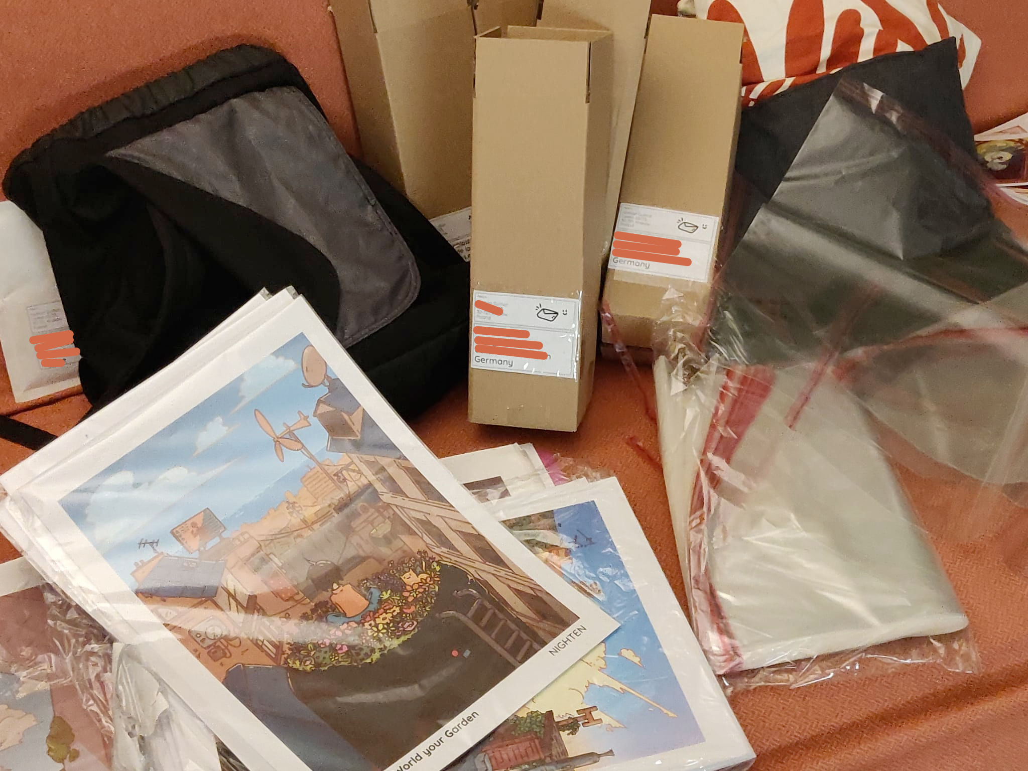 4 other cardboard box are on an orange couch, along with a black backpack, plastic sleeves and art prints of my illustrations