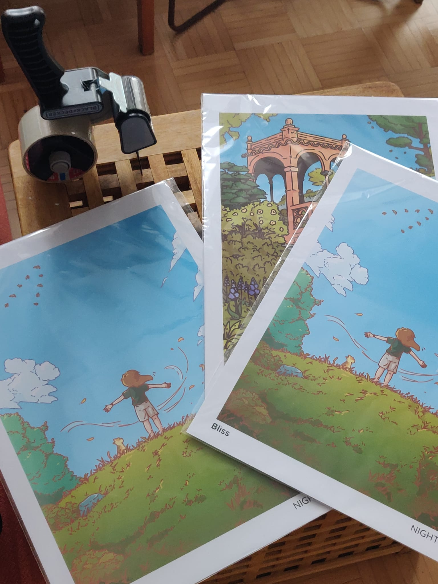 3 prints in plastic sleeves standing on a table, next to a big tape dispenser. 2 of the prints are identical and shows a woman with her arms spread on top of a hill, and the other one is of an old temple standing in a garden